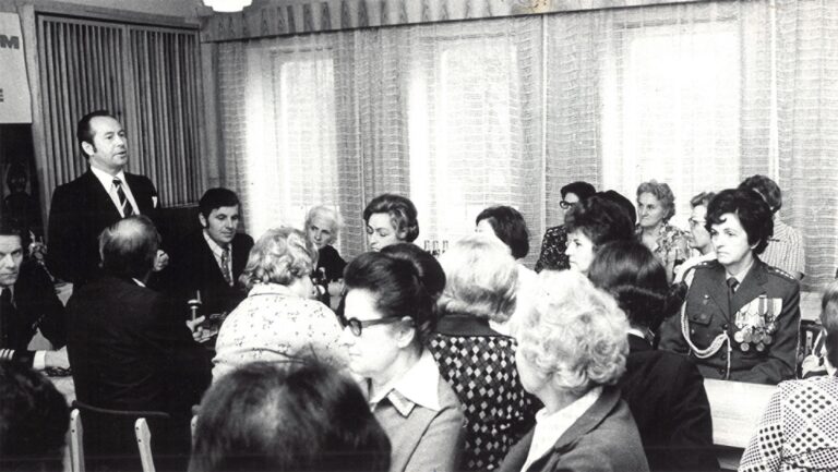 Czarno białe zdjęcie ze spotkania, na którym mężczyzna w garniturze przemawia do zgromadzonych kobiet